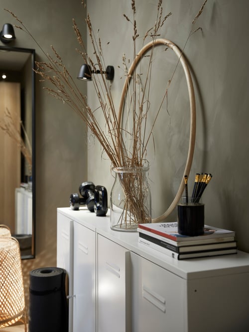 Side shot of a cabinet with drawers on which is a wreath, books, and a vase with twigs, with a wall lamp mounted on the wall.