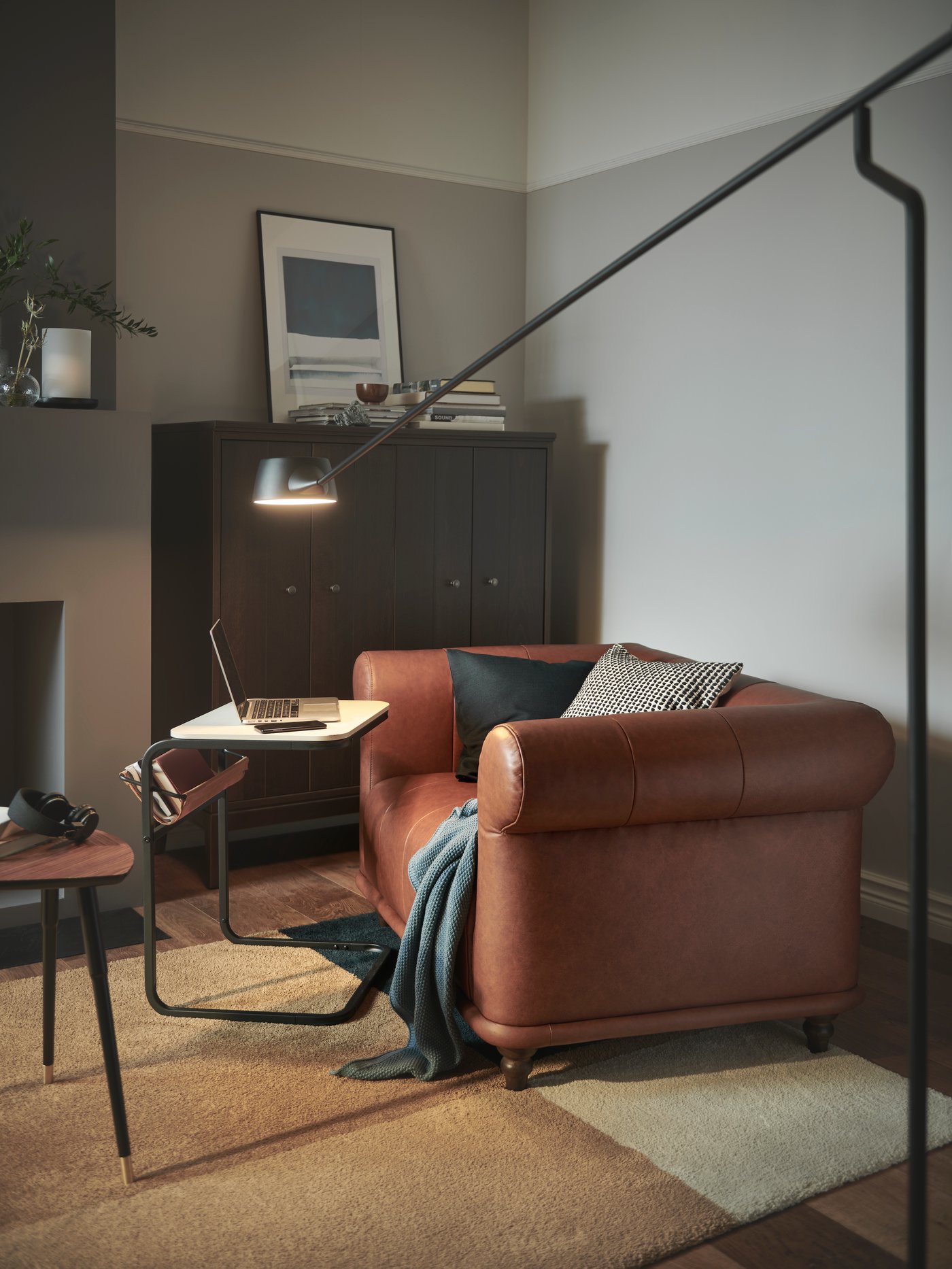 Zona de un salón con un sillón VISKAFORS en marrón, una lámpara de pie VÅGHÖJD encendida, un armario IDANÄS y una mesa auxiliar OLSERÖD.