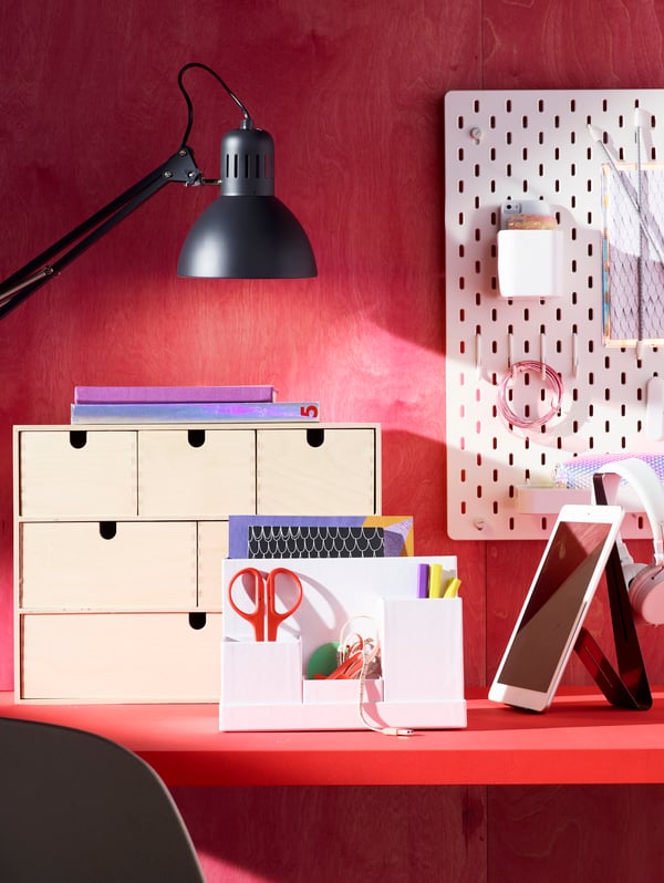 A desk in the color red with a red background that is filled with various IKEA products such as a lamp, notice board, drawers, scissors and pens.