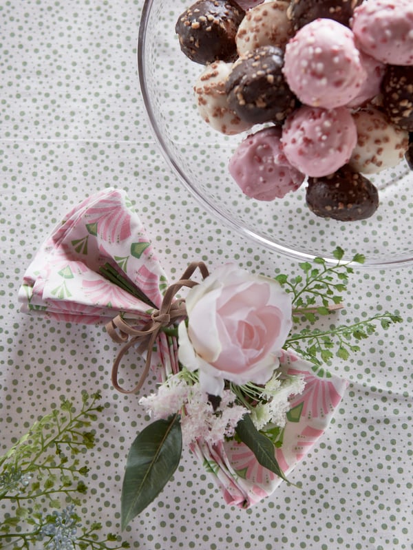 Una servilleta INBJUDEN decorada con una rosa artificial INBJUDEN de color rosa sobre una mesa junto a un cuenco de cristal lleno de dulces.