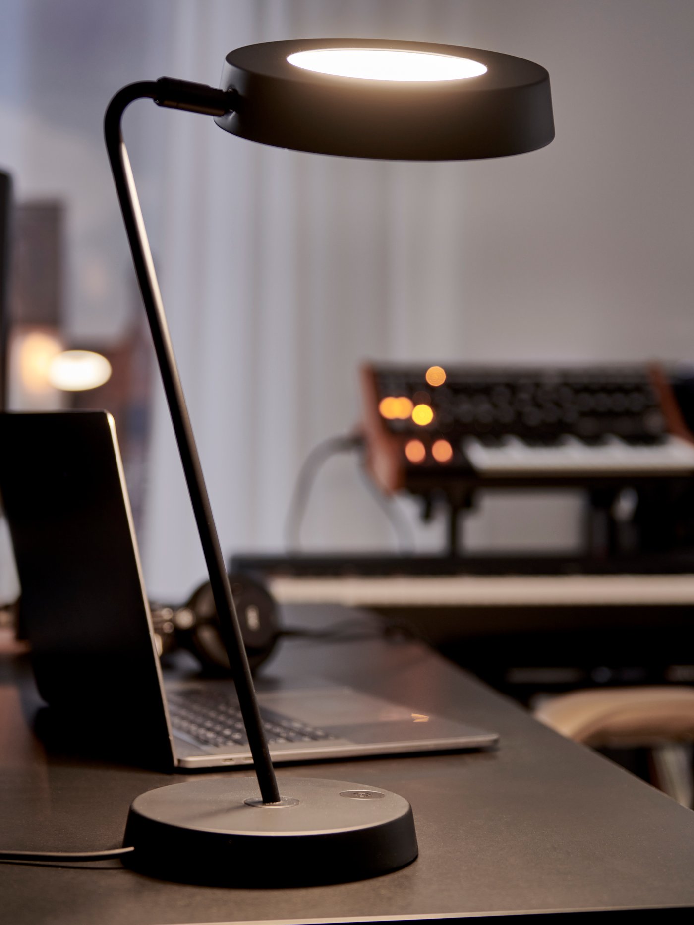 A lit black OBEGRÄNSAD LED work lamp on a black desk next to an open laptop with keyboards in the background.