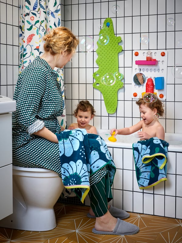 A mother is watching on as her kids play in the bath. She is holding a towel and behind her hangs different bath accessories.