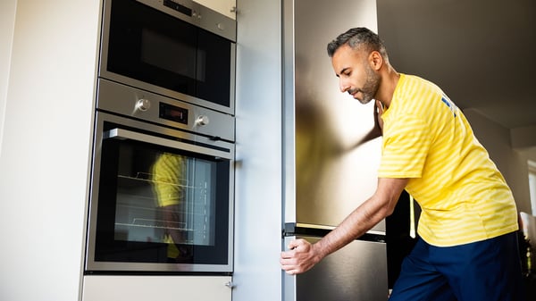 An IKEA co‑worker installs and adjusts a built‑in kitchen refrigerator beside integrated ovens in a modern home kitchen.