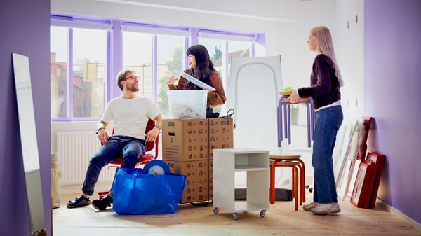 Three young people with lots of moving boxes, bags and furniture, obviously moving into a new room. Big windows in the back.