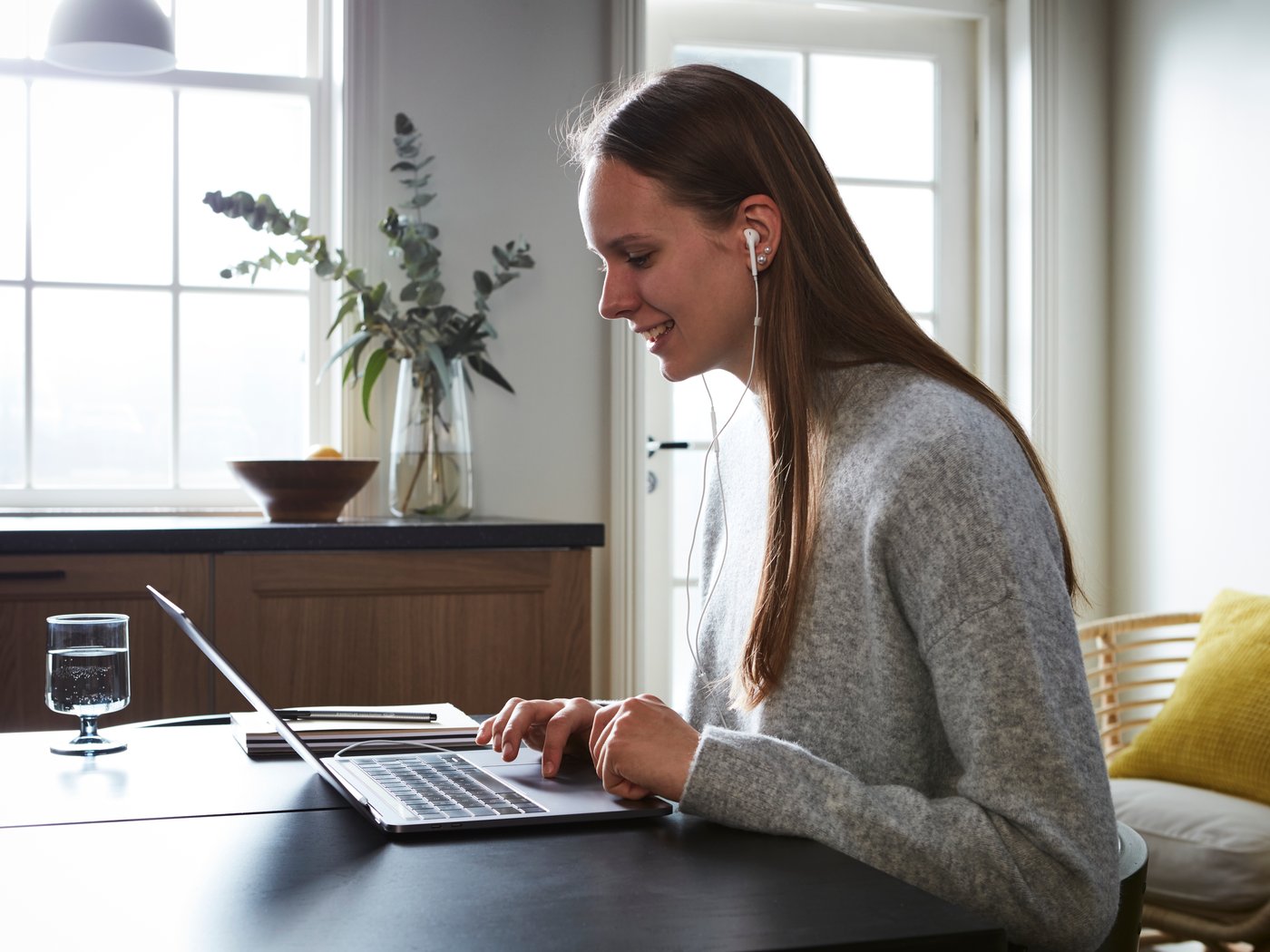Person am Tisch mit Laptop und Kopfhörern, Glas Wasser, Bücher und Pflanzen im Hintergrund bei Tageslicht.