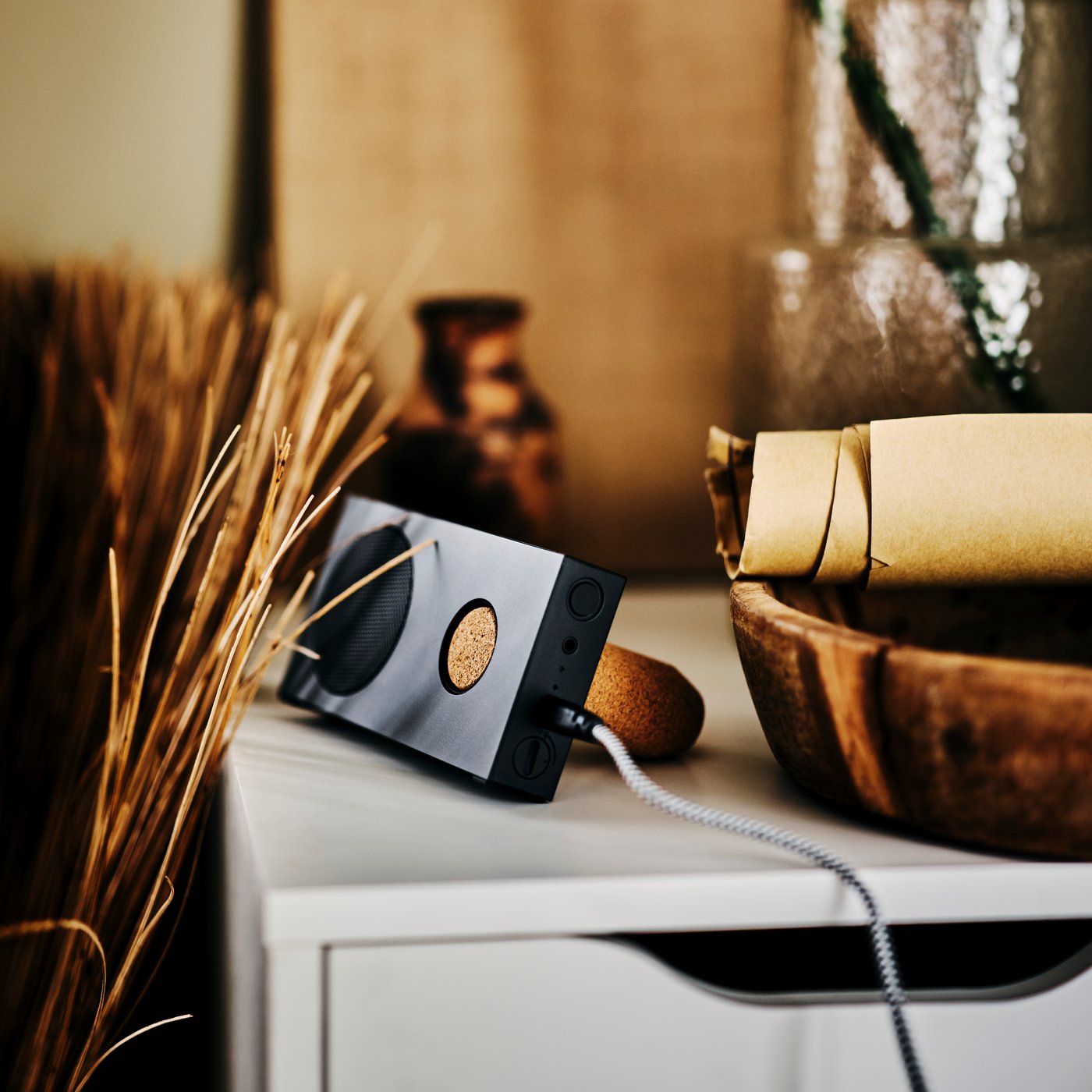 An ENEBY portable Bluetooth speaker on the corner of an ALEX drawer unit next to a wooden bowl holding a roll of brown paper.