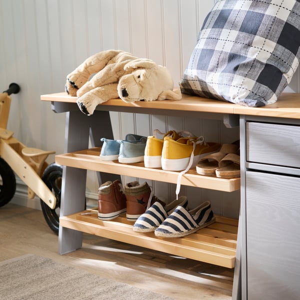 Close-up of KALLSÖ bench with shoe storage with a mix of shoes on the shelfs, and a soft toy and pillow on top.