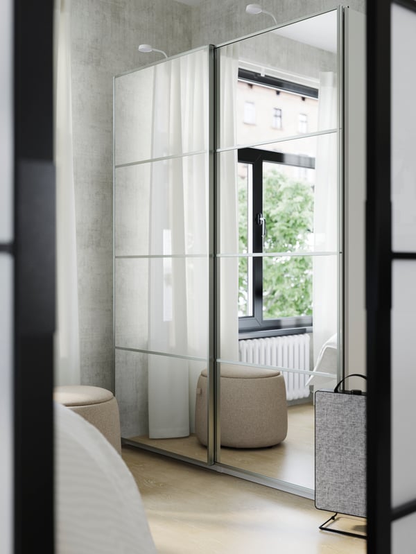 A window and sheer curtains are reflected in the closed, mirror glass sliding doors of a PAX/AULI wardobe on a bedroom wall.