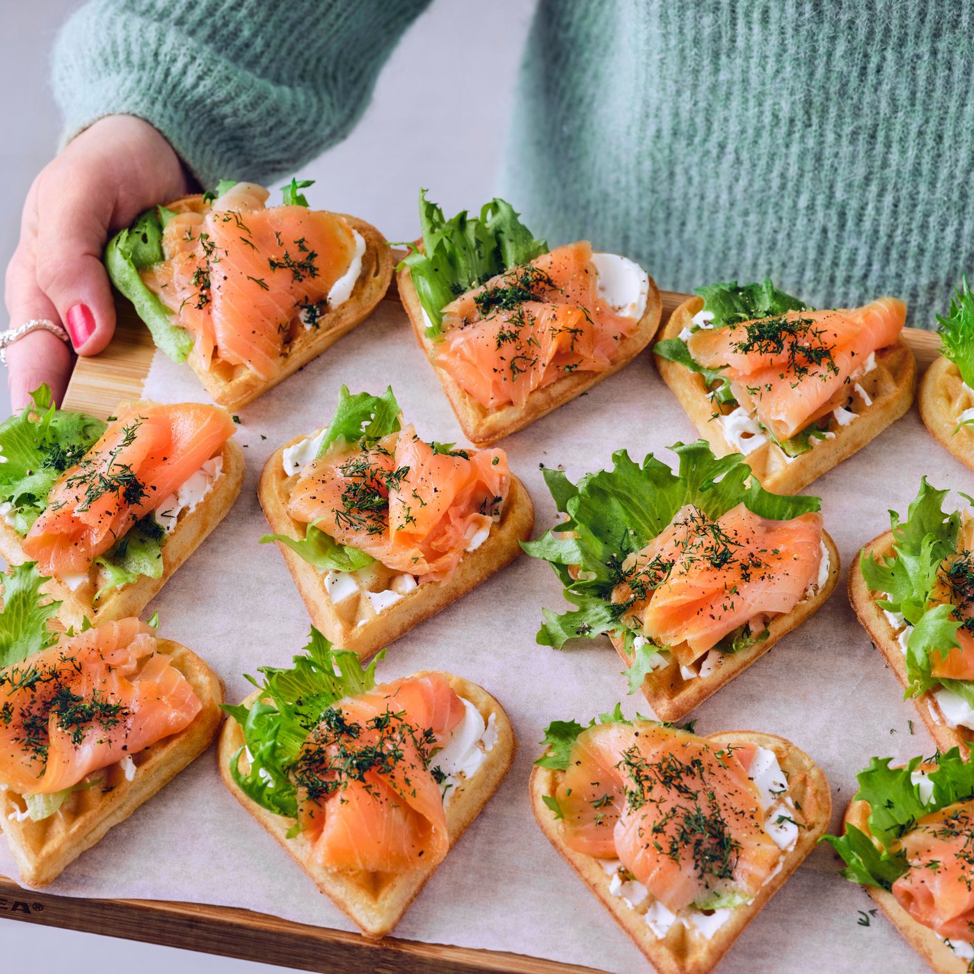A person in a blue sweater is holding a wooden board and it has heart-shaped waffles with salmon, cream cheese and lettuce.