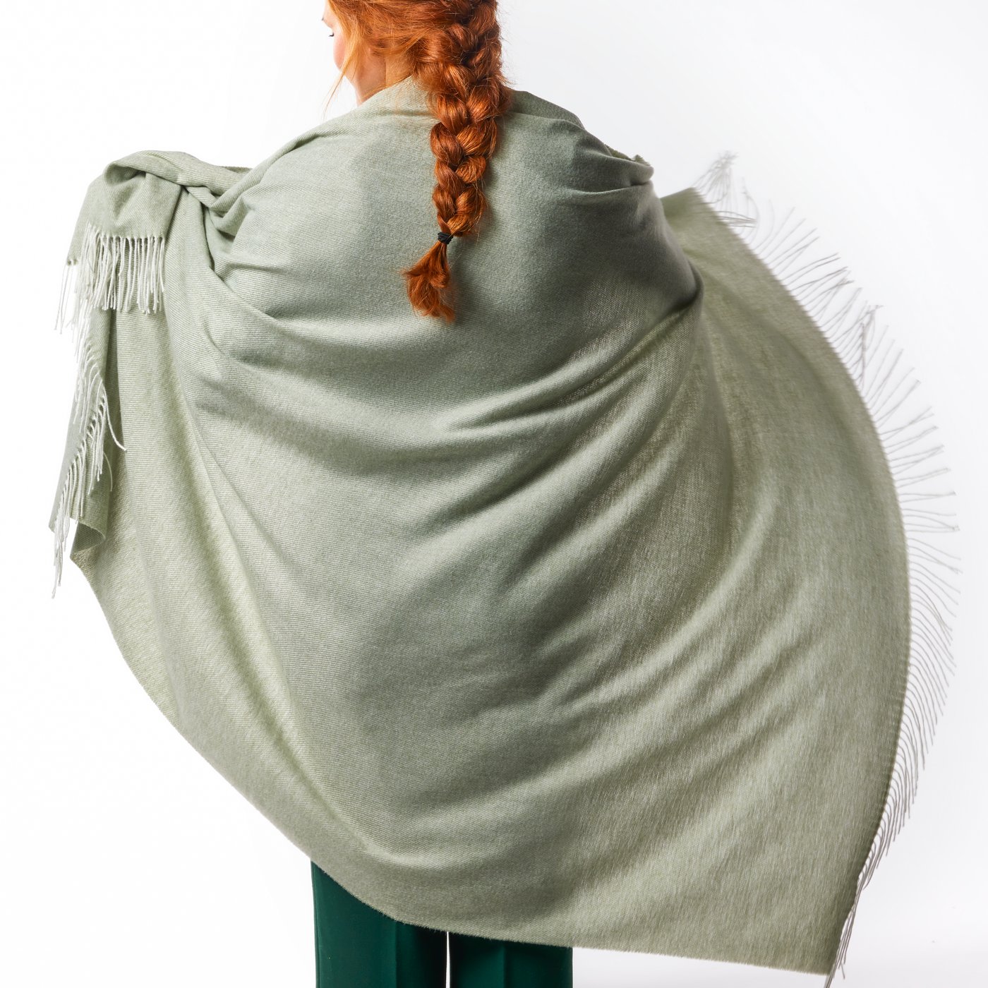 A person seen from behind has red hair in a long braid. They are wrapping a soft green HOLMVI throw around their back.