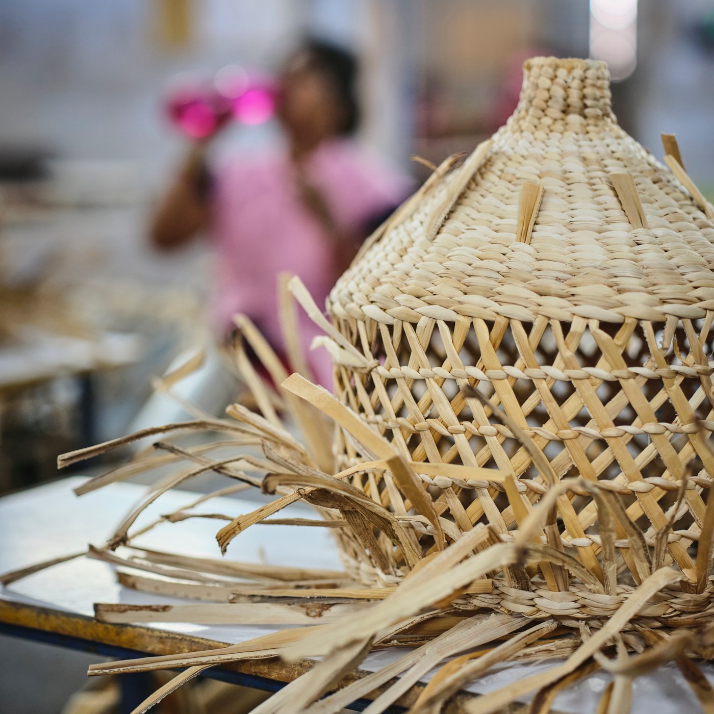 Un abajur de lustră MÄVINN pe jumătate lucrat este așezat pe o masă. Fâșiile din fibră de bananier ies în toate direcțiile.