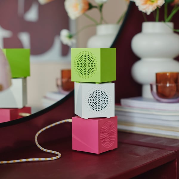 Three KALLSUP portable Bluetooth speakers in different colours stand on top of each other near a PÄRLHÄGG vase.