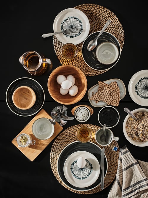 Top view of table with white and grey dinnerware, and plates on 2 place mats made of woven seagrass and black thread.