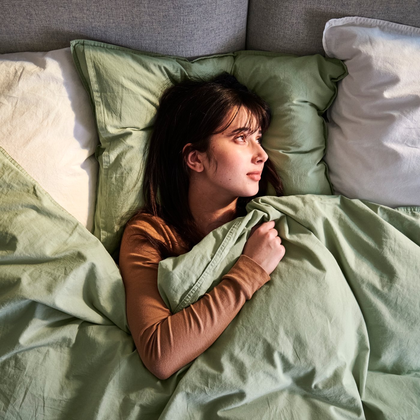 A person is on a sofa bed, looking off to the side, snuggled in a pale grey-green ÄNGSLILJA duvet cover and pillowcase.