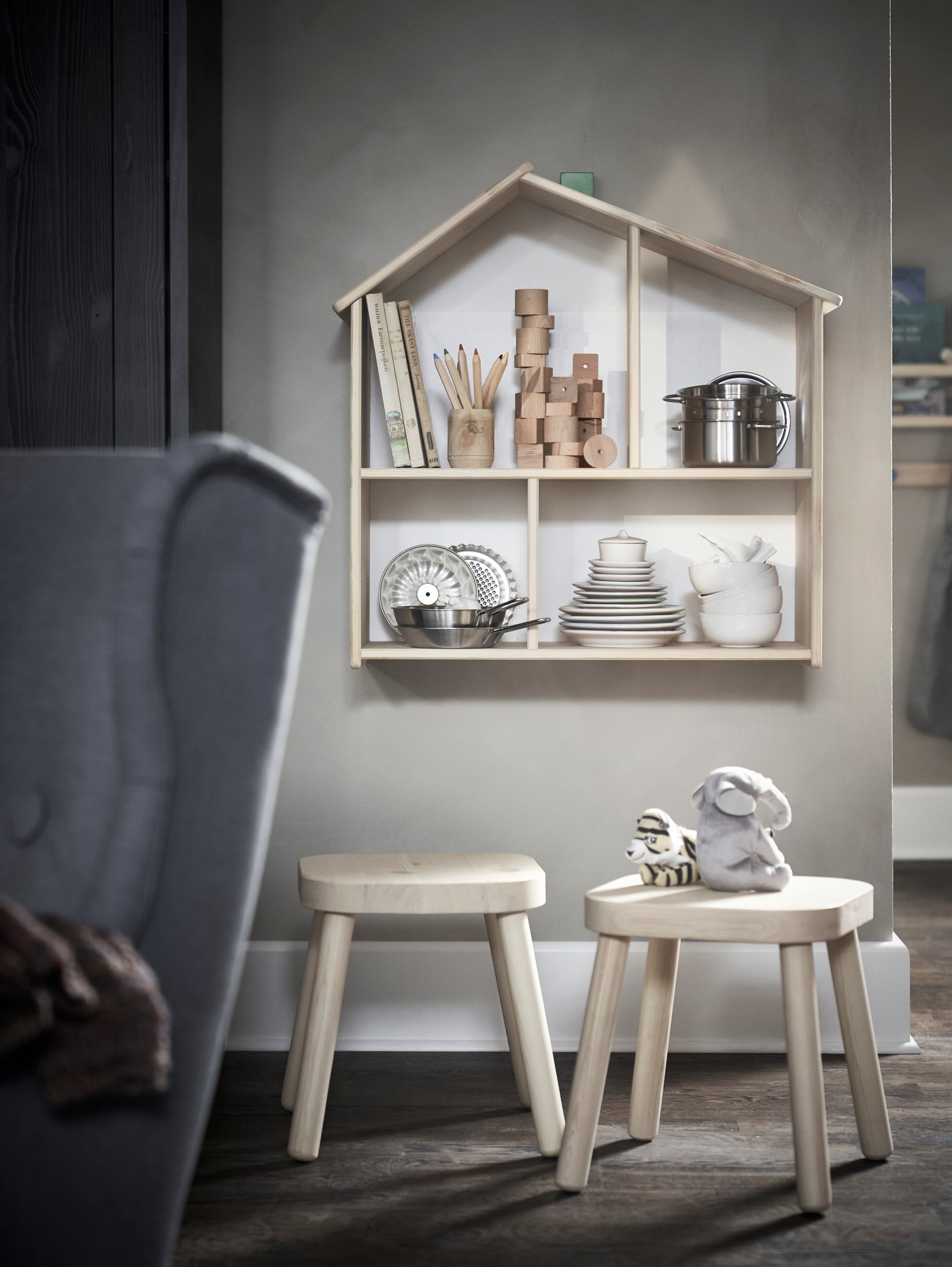 Dormitorio infantil con una casa de muñecas y estantería, un taburete y un sillón para niños, peluches, libros, ceras, ollas, sartenes y platos.
