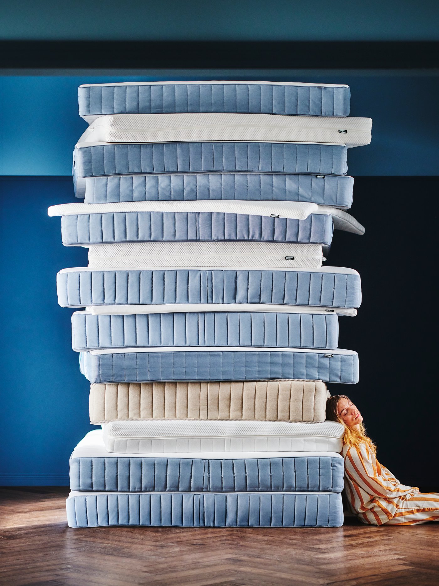 In a blue room, a person in striped pyjamas is sitting on the floor and leaning against a stack of IKEA mattresses.