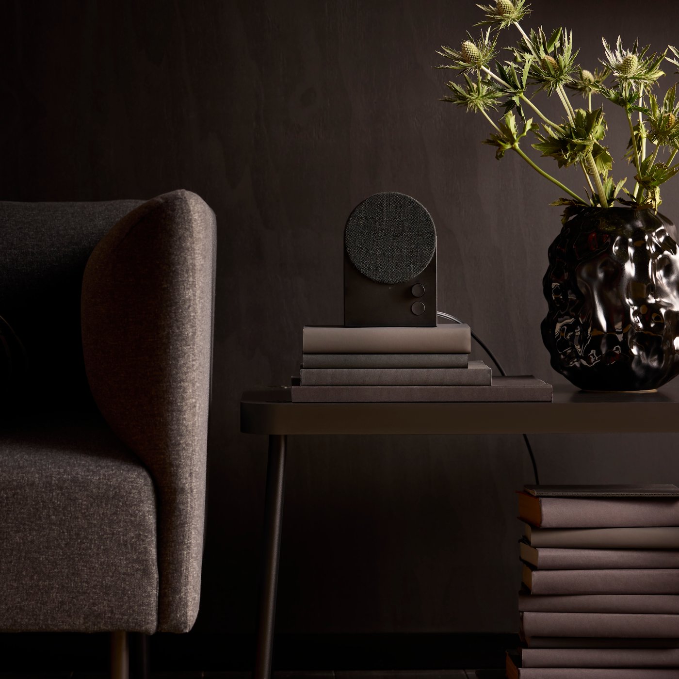 A black NATTBAD Bluetooth speaker sitting on a stack of books on a table beside a dark grey armchair.