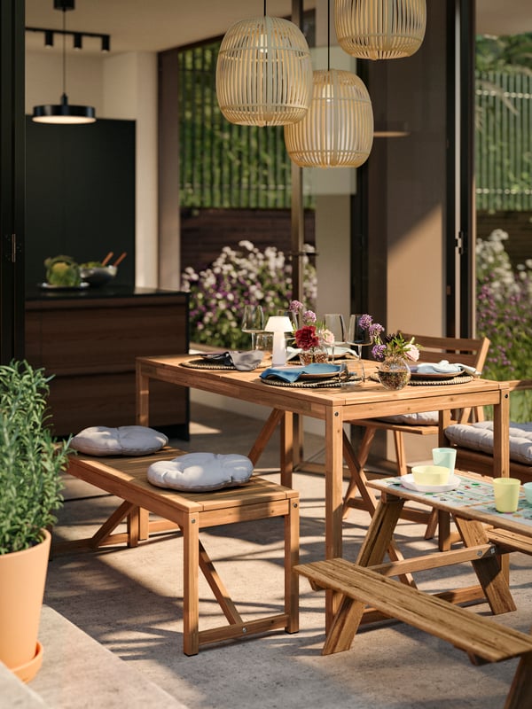 A patio dining area with light brown stained NÄMMARÖ outdoor furniture: a table, two chairs and a bench prepped for lunch.