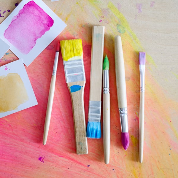 The 6 different sizes of MÅLA paint brushes, all dipped in different colours, laying on a wooden base.