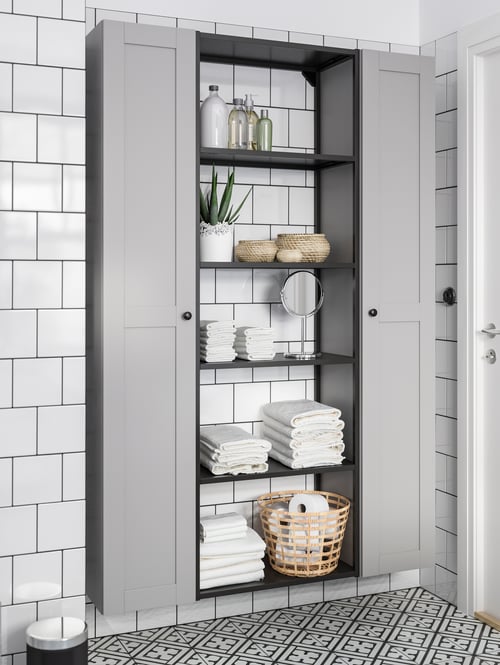 Two high cabinets surround a high frame with shelves on which are boxes with lid, folded towels, and a round mirror.