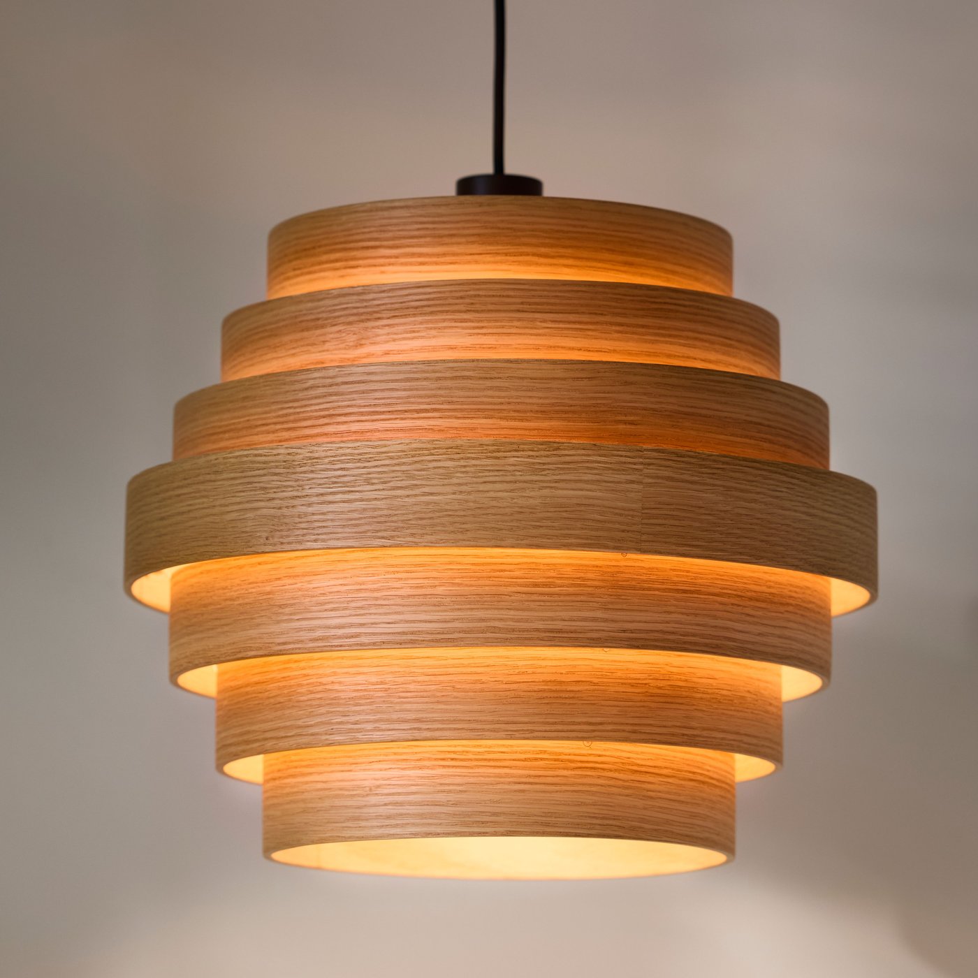 A white stained oak veneer DYKARKLOCKA pendant lampshade with a lit light bulb inside it hangs from a ceiling.