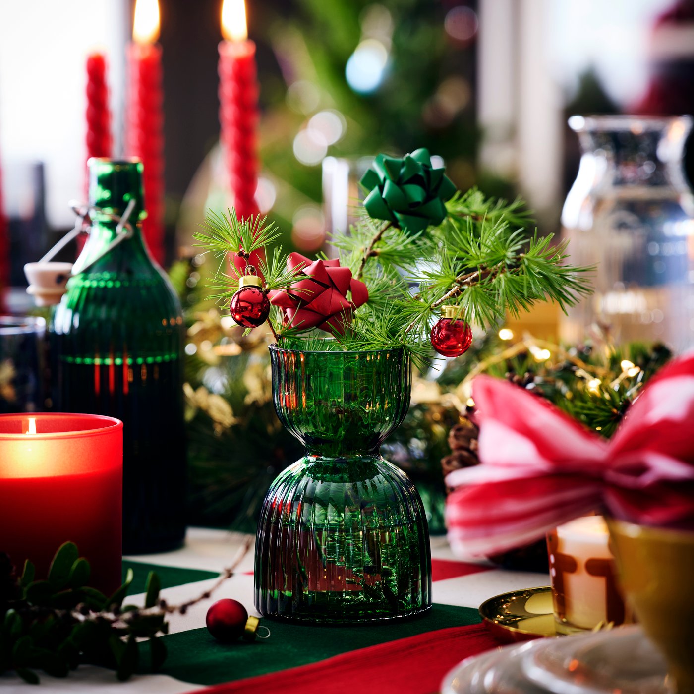 Frans: Une table décorée de façon festive avec des vases en verre vert contenant des branches de sapin et des ornements rouges, entourée de bougies et de décorations de Noël.