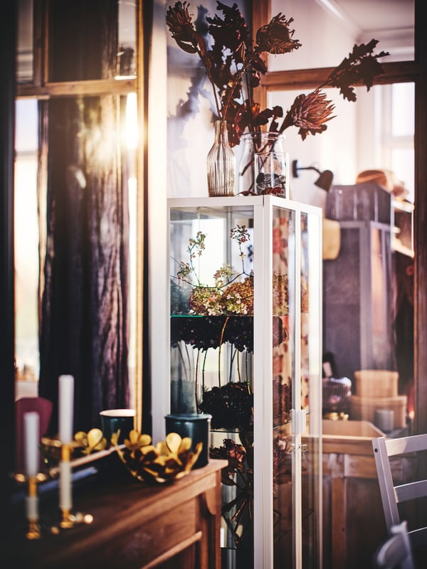 A MILSBO glass-door cabinet containing vases with flowers within and on top. A STOCKHOLM bowl is on a surface to the side.