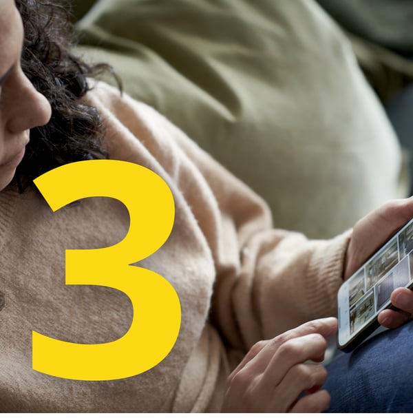 Kobieta z kręconymi włosami w beżowym swetrze przegląda zdjęcia na smartfonie, oparta o zielone poduszki. Żółta cyfra 3 obok.