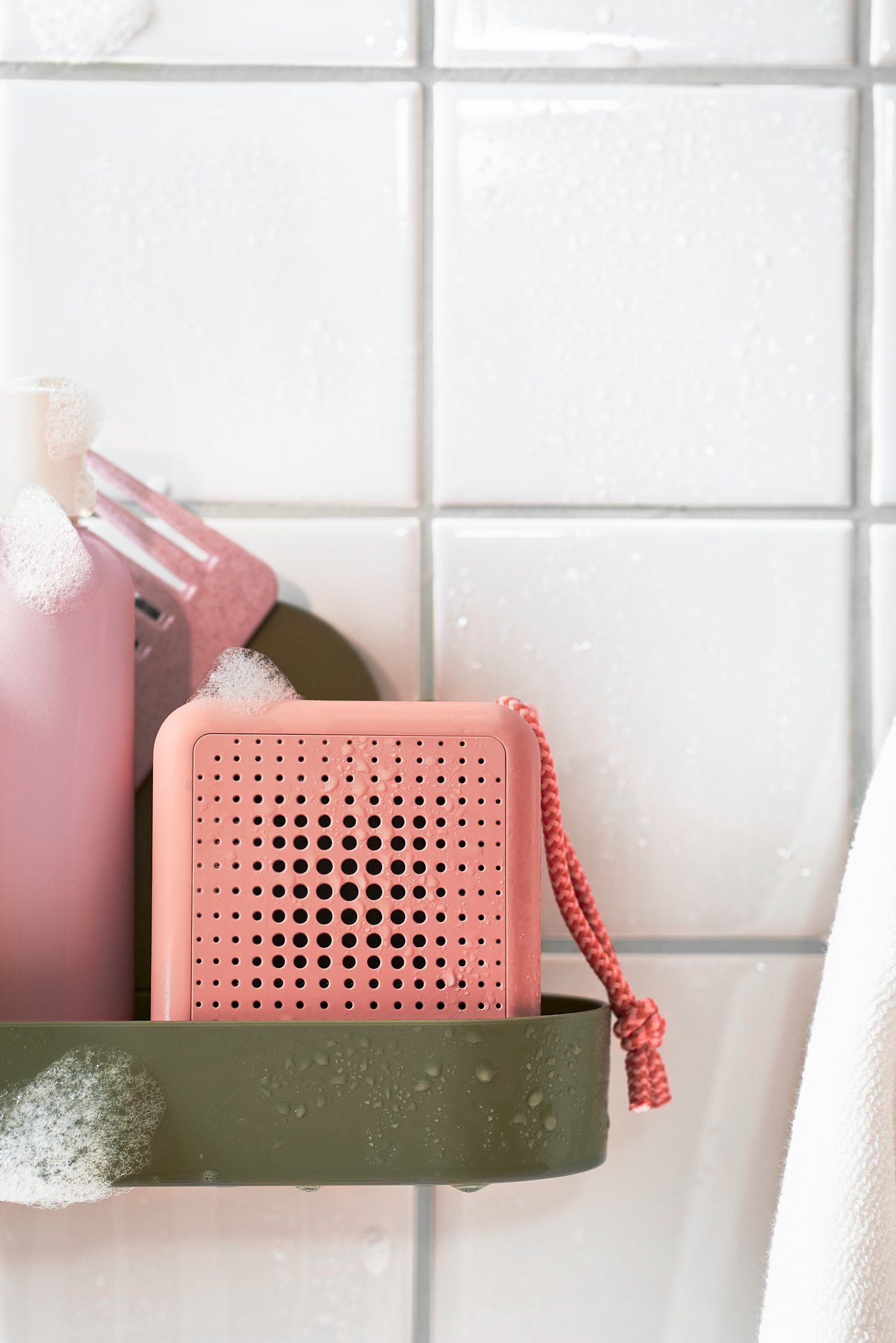 Ein wasserdichter, rosafarbener tragbarer Bluetooth-Lautsprecher der Marke VAPPEBY steht in der Dusche auf einem graugrünen ÖBONÄS-Wandregal mit Saugnapf.
