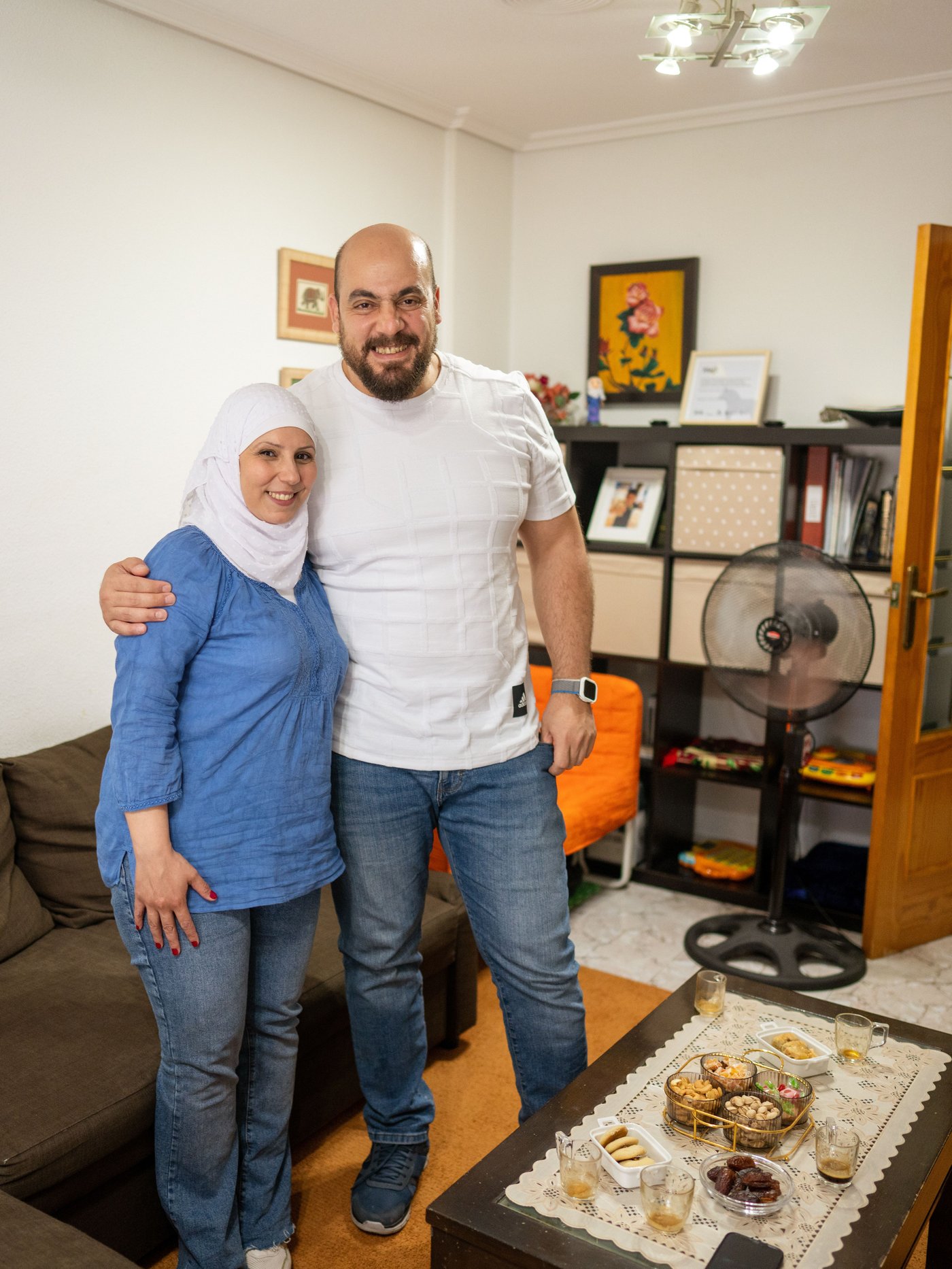 A couple stands in their living room, holding each other while smiling at the camera before having a coffe break.