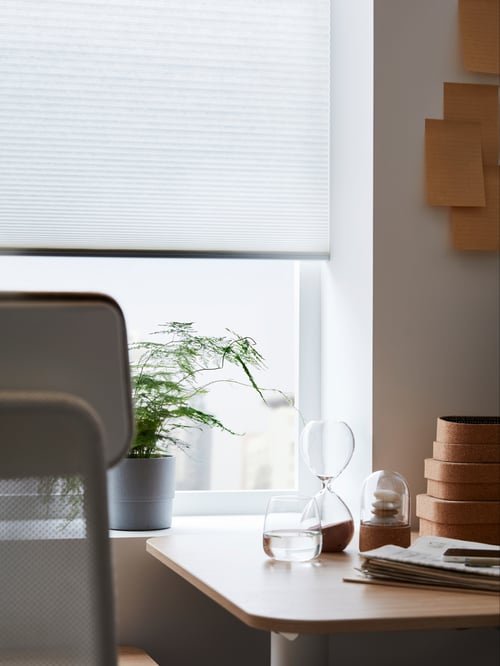 A white HOPPVALS cellular blind with a honeycomb structure halfway up a window, above a potted plant by a work desk and chair.