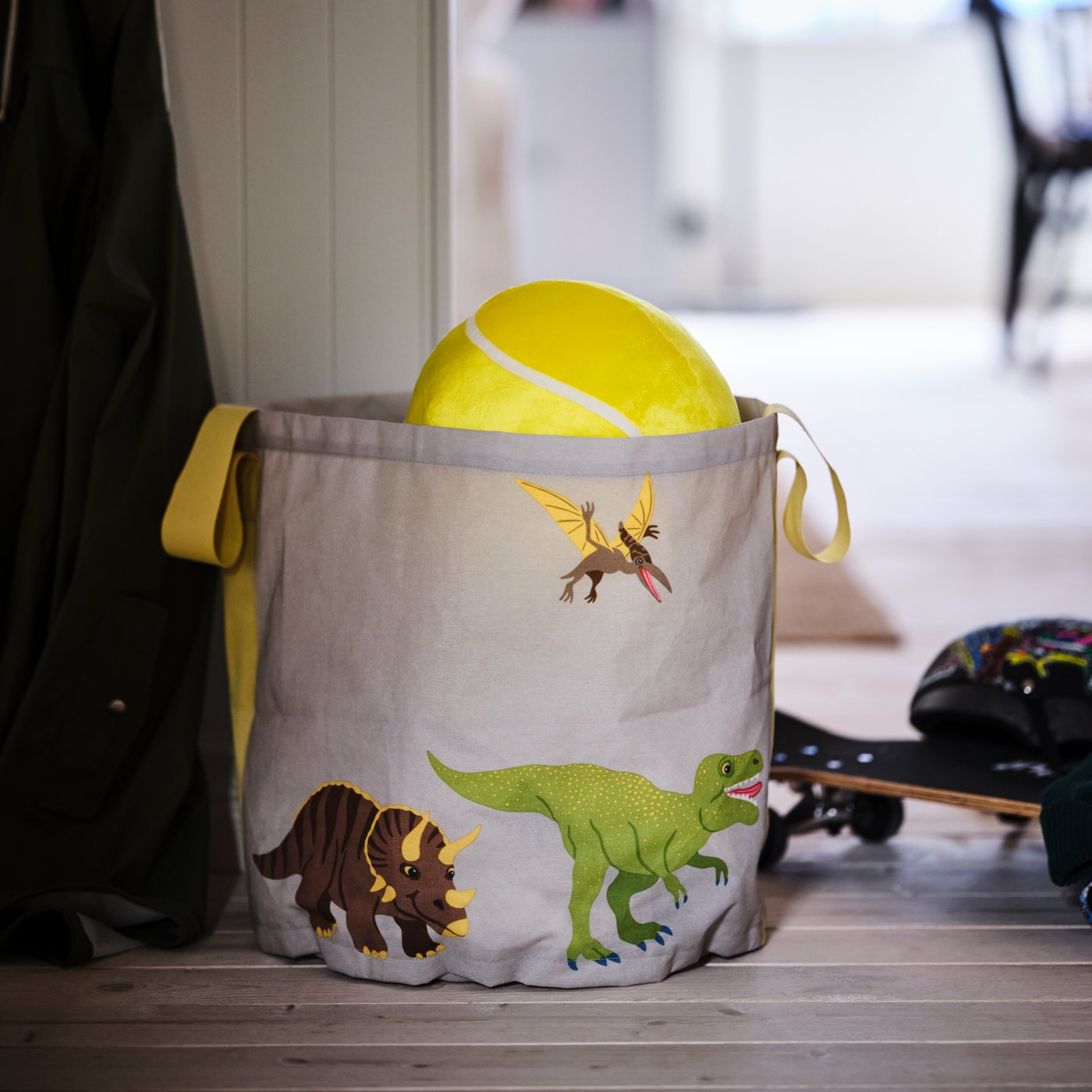 A JÄTTELIK dinosaur-decor storage bag with toys in and around it standing on the wooden floor of a hallway.