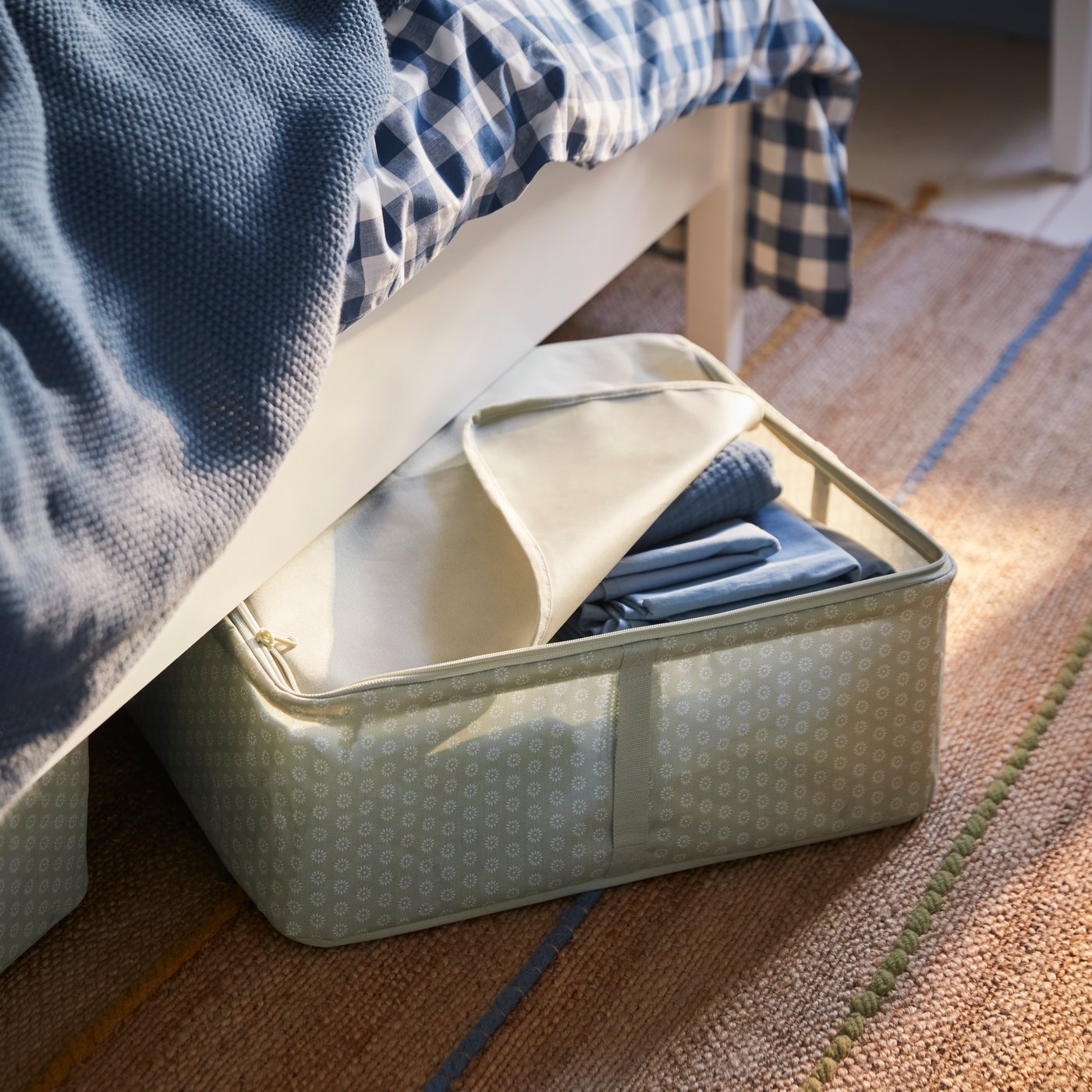 Una caja HEMMAFIXARE en gris-verde pálido llena de ropa de cama azul que sobresale de debajo de una cama blanca.