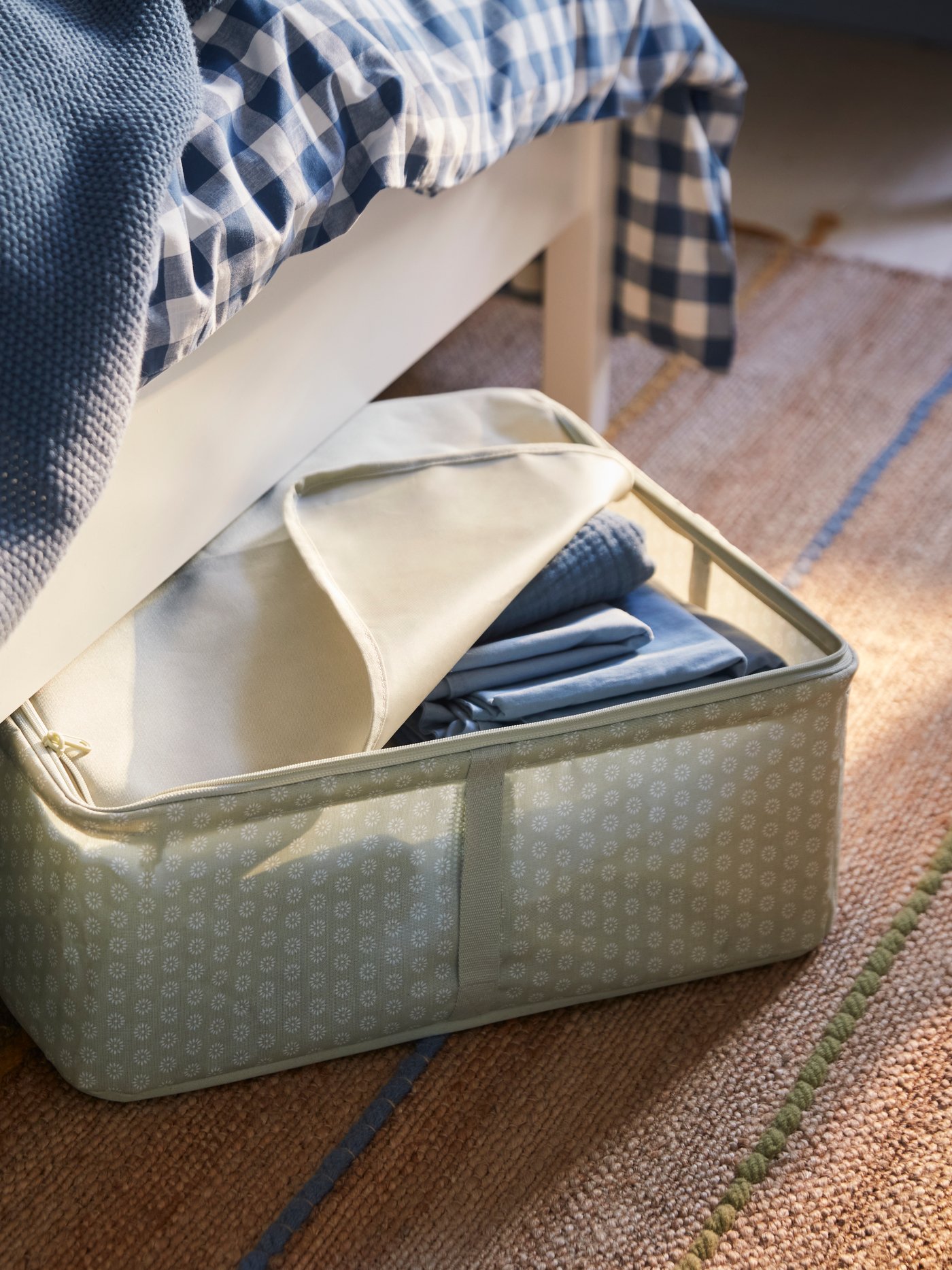 A pale grey-green HEMMAFIXARE storage box filled with blue bedding sticks out from under a white bed.