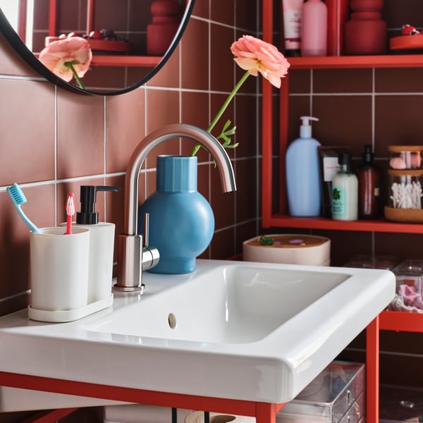 Weißes rechteckiges Becken mit silberner Armatur, Zahnbürstenhalter, blauer Vase mit Blumen und roten Regalen – gut organisierter Badezimmerplatz mit Pflegeprodukten.