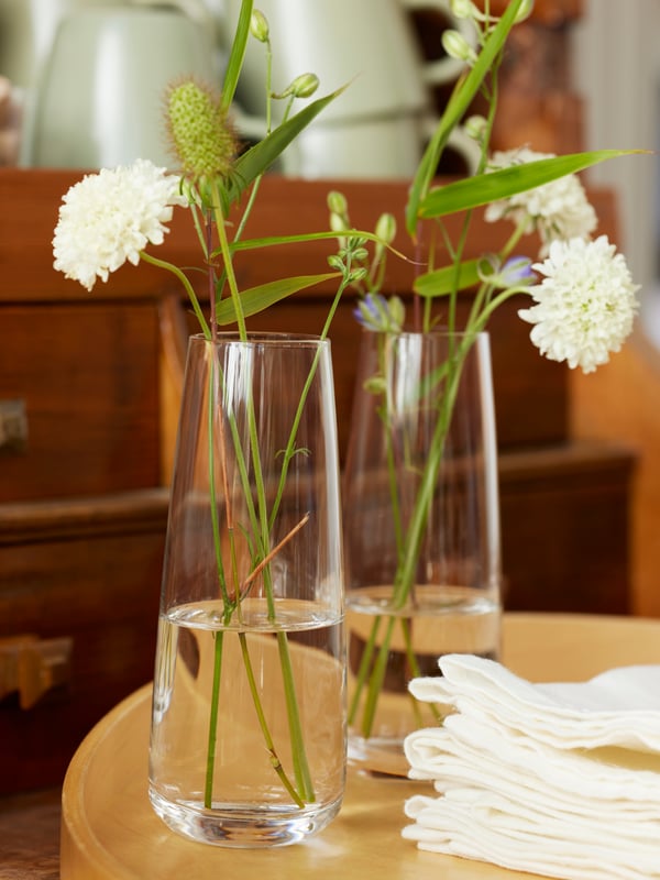 On a light and beautiful table setting with two white plates filled with sallad is a BERÄKNA glass vas with flowers.
