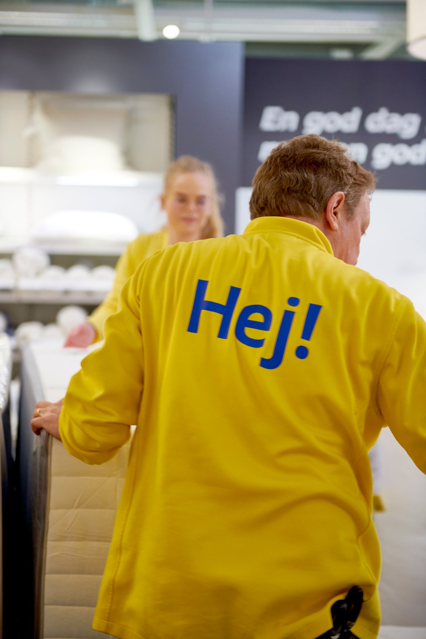 Keltaiseen työasuun pukeutunut IKEA-työntekijä, jonka paidan selässä on sana ”Hej!”.