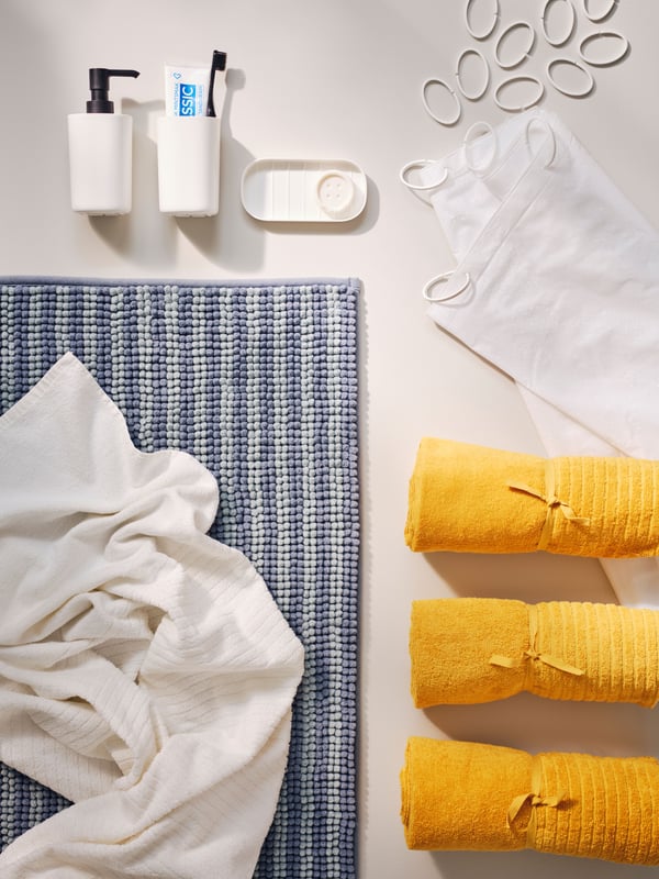 An image showing a neatly organized set of bathroom items on a light surface. The items include three rolled yellow towels, a white face mask, two soap dispensers, a bar of soap in its dish.