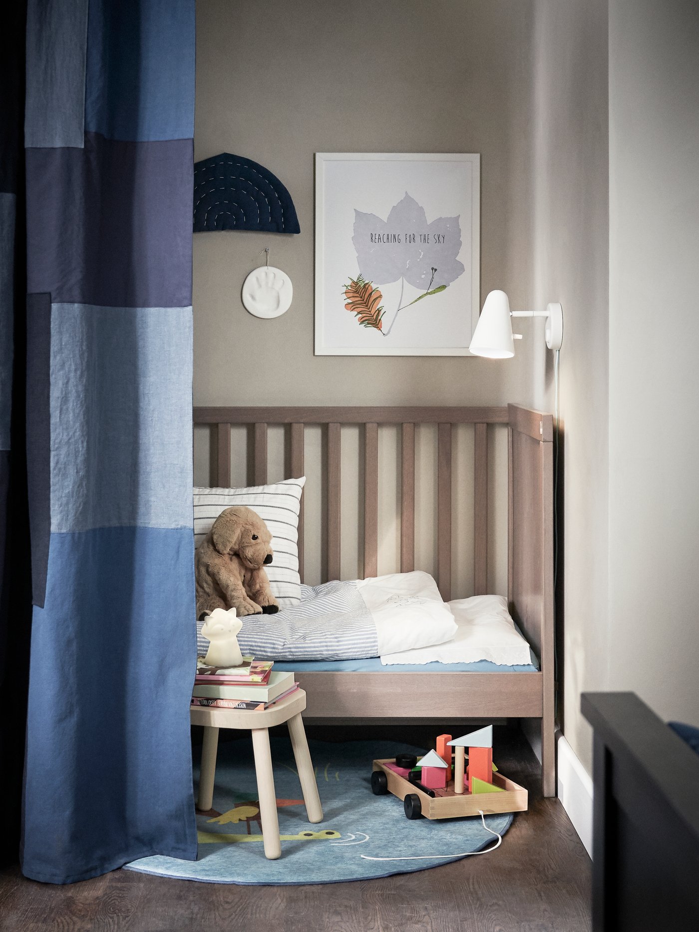 Gemütliche Kinderecke der SUNDVIK Serie mit einem kleinen Holzbett, einer weißen Bettdecke mit blauem Rand und einem gestreiften Kissen. Ein brauner Plüschbär sitzt auf dem Bett. Daneben steht ein kleiner Holzhocker mit Kinderbüchern. 