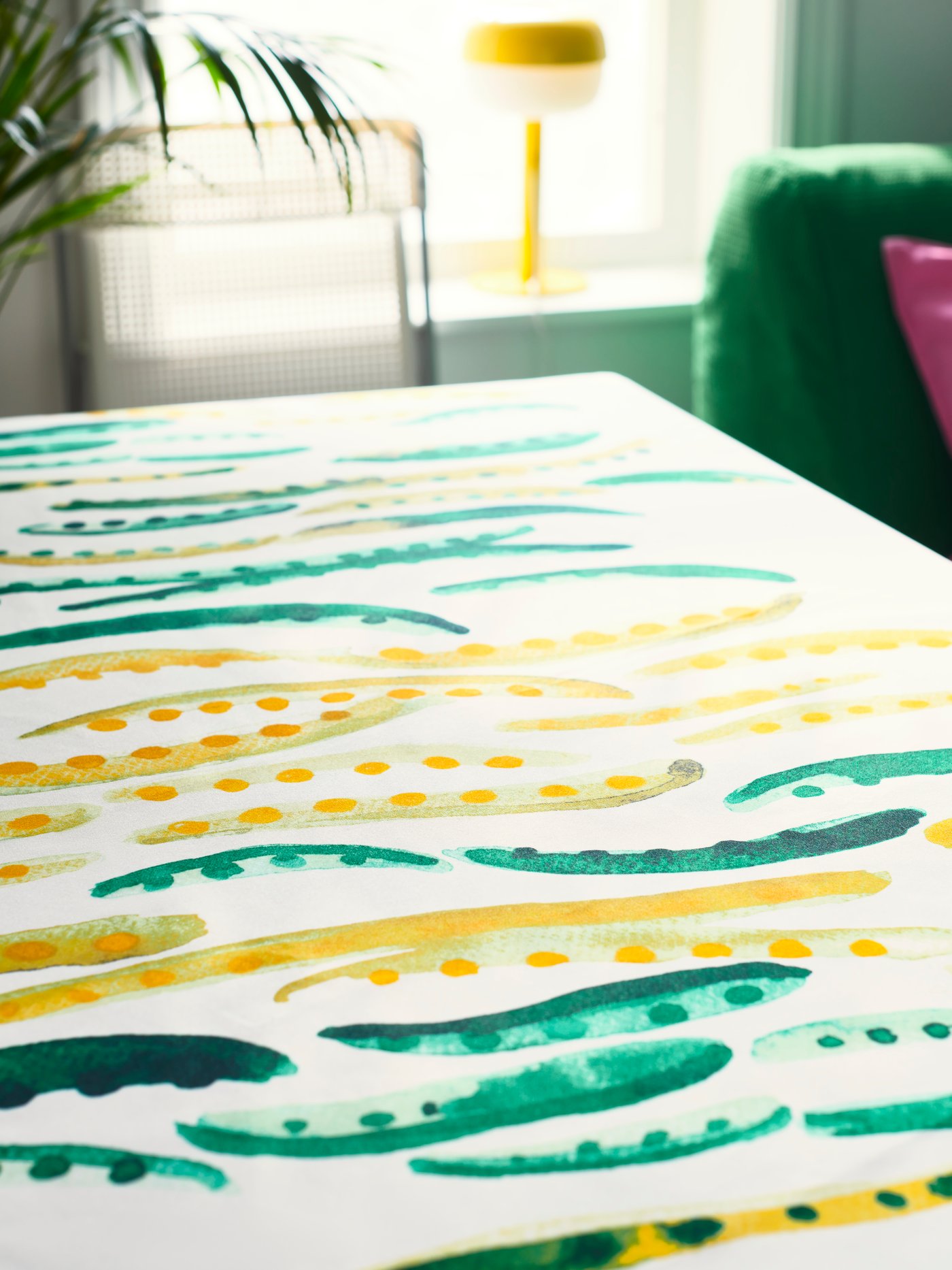 A tablecloth with yellow and green pea pattern is laid on a flat surface. A table lamp and plant are seen in the background.