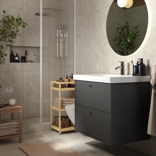 A sunlit zen bathroom with a dark-grey HAVBÄCK drawer and furniture in natural bamboo. A large green plant in the corner.