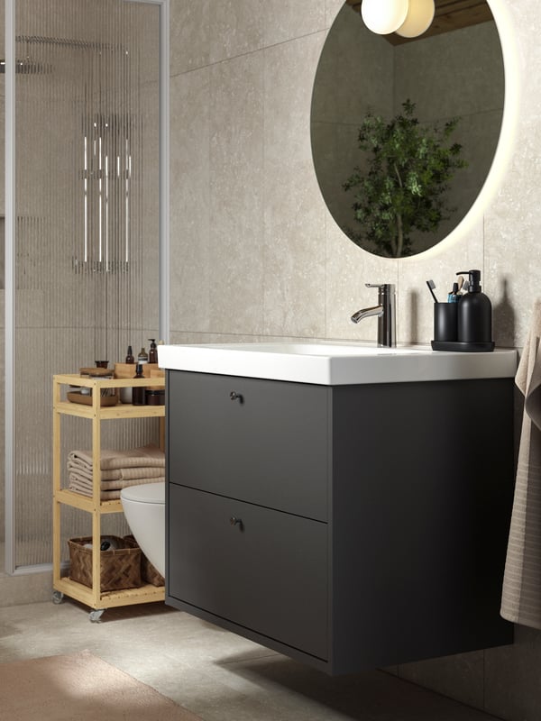 Salle de bain ensoleillée avec meuble à tiroirs HAVBÄCK gris foncé et meubles en bambou naturel. Une grande plante agrémente un angle de la pièce.