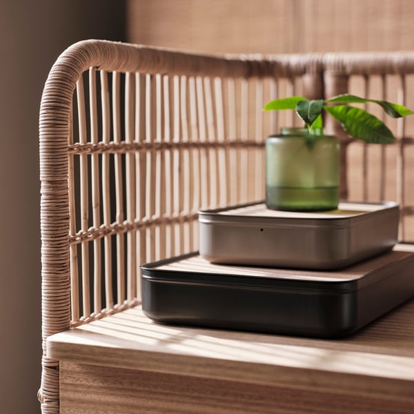 A corner of the FRYSKÅS rattan desk with two HARVMATTA boxes with lid on top, one in anthracite and one in dark grey-beige.