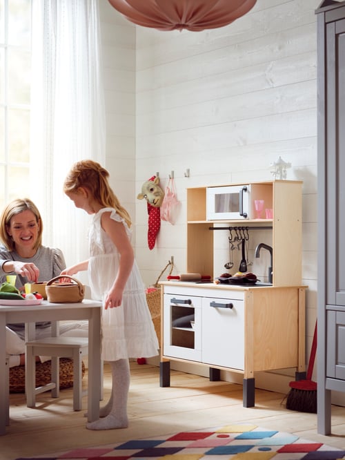 Une fille debout à une table d’enfants joue avec sa mère avec une corbeille de fruits, à côté d’une minicuisine DUKTIG.