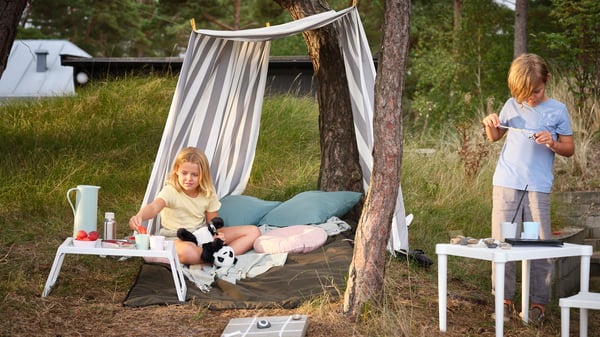 Two children outside, one eating a watermelon while seated under a tent and the other painting a pine cone.