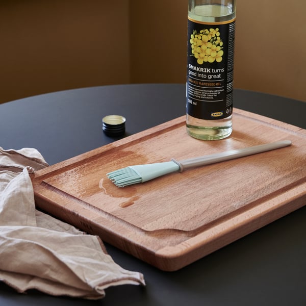 A PROPPMÄTT chopping board in beach with a small brush and a bottle of rapeseed oil on a table beside a cloth.
