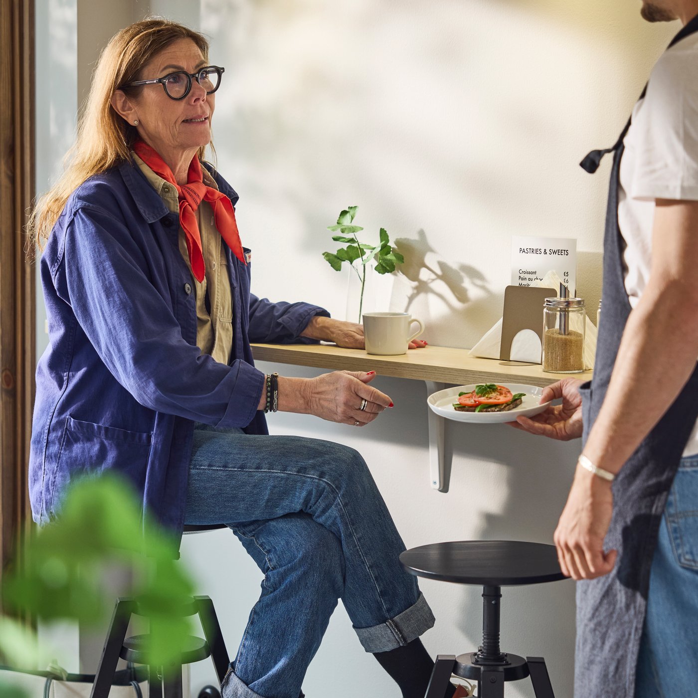 Kund som sitter vid cafédisken får en tallrik smörgås från baristan; kopp, växt och tillbehör på bordet i en ljus miljö.