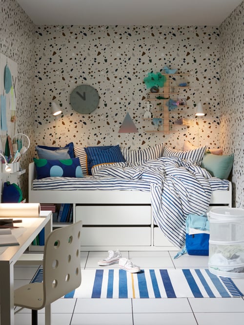 Weißes SLÄKT Bett in einem Raum mit gefleckter Tapete, einer Uhr, zwei Lampen und Regalen an der Wand, gestreifter Bettwäsche und einem Schreibtisch.