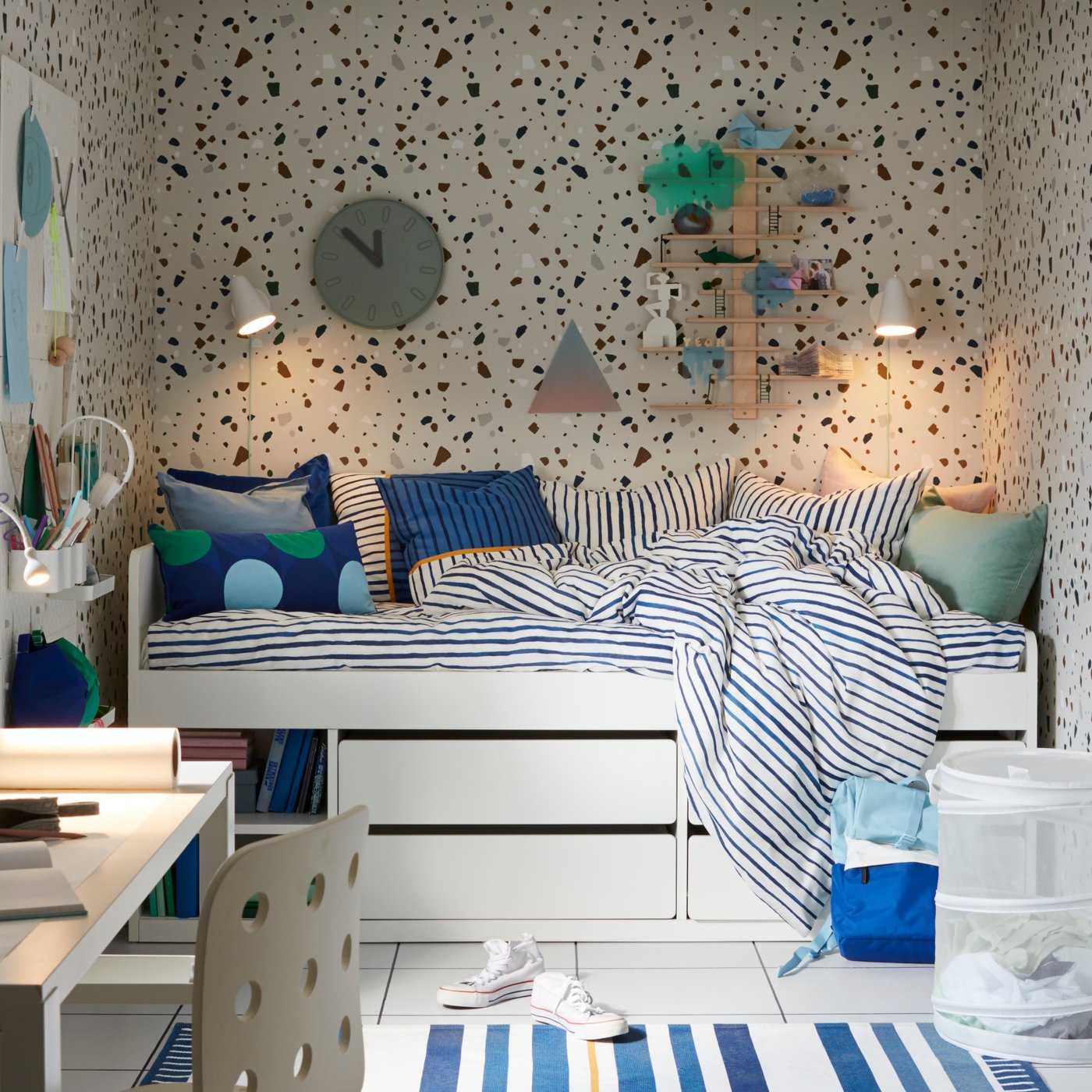 White SLÄKT bed in a room with speckled wallpaper, a clock, two lamps and shelves on the wall, striped bed linen and a desk.