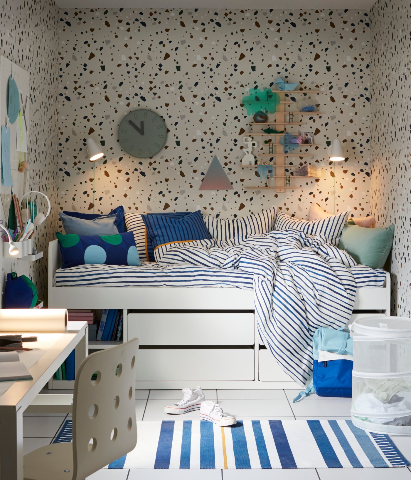 White SLÄKT bed in a room with speckled wallpaper, a clock, two lamps and shelves on the wall, striped bed linen and a desk.